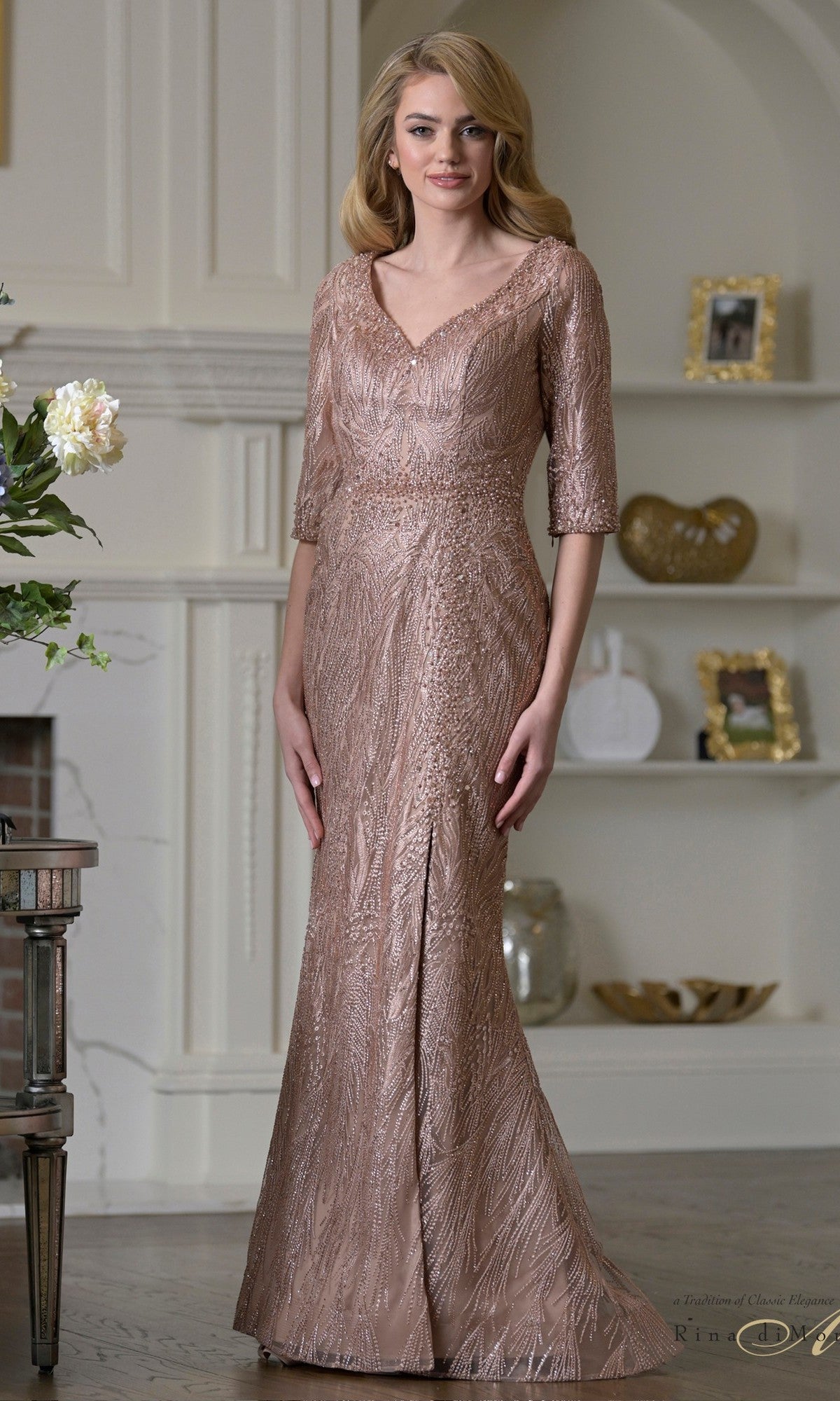 A woman wears the Long Formal Dress: Rina Di Montella RD3530 in rose gold, featuring embroidered mesh half sleeves, standing in a stylish room adorned with shelves and framed photos.